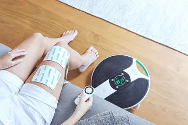 Image of a ladys legs with the thigh pads on her thigh and the medic knee to the side of her feet with the remote in her hand